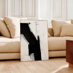 Bold black brushstrokes on cream canvas leaning against beige sofa in minimalist living room.