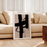 Minimalist black brushstroke art in white frame against beige sofa and wooden floor