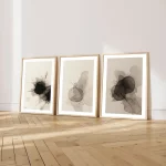 Three gold-framed abstract ink artworks leaning against white wall on wooden floor.