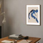 Minimalist blue brushstroke art above wooden desk with dried plants and design books.