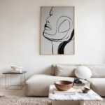 Abstract face line drawing displayed above cream sofa in minimalist living room setting.