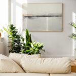 Abstract landscape painting above white sofa surrounded by houseplants in bright living room.