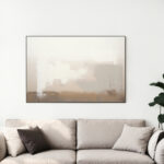 Abstract beige artwork in thin black frame hanging above neutral sofa with decorative pillows.