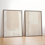 Two minimalist abstract artworks in neutral tones with black frames leaning against white wall.