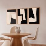 Minimalist dining area with black and cream abstract art trio above wooden table.