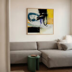Bold abstract painting featuring blue, black and yellow over modern gray sectional sofa.