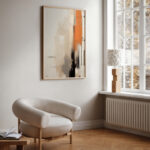 Abstract artwork in neutral tones with orange accents above beige curved armchair.