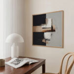 Geometric abstract artwork in earthy tones hangs above wooden desk with lamp and book.
