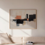 Abstract geometric artwork with black and orange shapes above beige sofa in sunlit room.