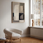 Abstract neutral wall art above cream armchair in minimalist sunlit living room.