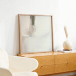 Abstract artwork in wooden frame on oak credenza with minimalist decor and white vase.