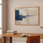 Abstract navy and beige wall art above wooden desk with books in minimalist space.