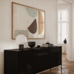 Abstract artwork in earthy tones over black credenza with lamp and decorative objects.