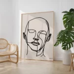 Minimalist line art portrait in wooden frame beside monstera plant and rattan chair.