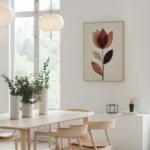 Framed abstract flower art print with warm earthy tones, displayed on a light wall above a wooden dining table with chairs and green plants, in a bright, minimalist dining area.