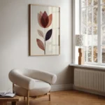 Framed abstract flower art print with reddish-brown and purple translucent shapes, hung on a light wall next to a large window, a radiator, and a light-colored armchair in a cozy nook.