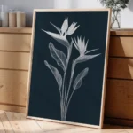 Framed white line art print of Bird of Paradise flowers on a dark background, leaning against a light wall on a wooden dresser with a glass vase and decorative items.
