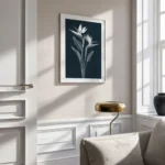 Framed white line art print of Bird of Paradise flowers on a dark navy background, displayed on a light wall with decorative wainscoting and a small table lamp below.
