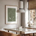 Framed botanical illustration of a white-flowered plant on a muted olive green background, hung on a wall above a modern dining table with a pendant light in a home.
