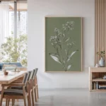 Framed botanical illustration of a white-flowered plant on a muted olive green background, hung on a wall in a bright modern dining room with wooden furniture.