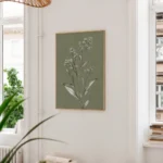 Framed botanical illustration of a white-flowered plant on an olive green background, hung on a light wall next to a large window and a tall green plant in a bright room.