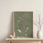 Framed botanical illustration of a white-flowered plant on a muted olive green background, placed on a light wooden table or console with a white vase and dried branches.