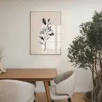Framed black and white botanical branch art print, hung on a neutral wall in a contemporary dining area with a wooden table, chairs, and a large potted olive tree.