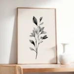 Framed black and white botanical branch art print, leaning on a light wooden dresser or sideboard next to a white vase in a neutral-toned room.