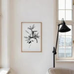 A vertical framed art print featuring an intricate black and white line drawing of a leafy branch, hanging on a clean white wall. To its right is a tall black floor lamp and a large window with multiple panes, letting in bright natural light. Part of a light-colored armchair is visible in the foreground.