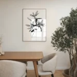 Framed black and white line art print of a leafy branch, displayed on a neutral wall in a contemporary dining room with a wooden table, chairs, and a large potted olive tree.