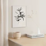 Framed black and white line art print of a leafy sprig, hung above a light wooden shelf or side table with a minimalist vase and books, bathed in warm natural light.