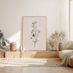 Vertical framed art print of a minimalist black and white botanical sprig on a blush background, leaning on the wooden floor in a serene, sunlit bedroom with neutral tones.