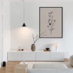 Black framed black and white botanical sprig art print on a blush background, displayed above a sleek white console table with a minimalist vase and black pendant light.