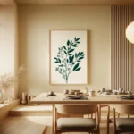 Framed art print showcasing a dark green botanical sprig on a light background, hung on a light wall in a modern dining room with a wooden table and chairs.