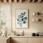 Framed art print of a dark green botanical sprig on a light background, adorning a light wall above open wooden kitchen shelves and a countertop with minimalist decor.