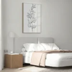 Framed black and white botanical sprig line art print, hung on a light gray wall above a bed with white bedding and a wooden bedside table in a modern bedroom.