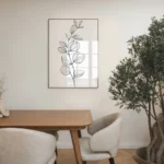 Framed black and white botanical sprig line art print, hung on a neutral wall above a wooden dining table with chairs and a large potted olive tree in a contemporary dining area.