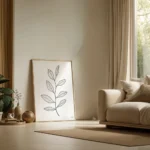 Large framed black and white line art print of a delicate botanical sprig, leaning against a neutral wall next to a beige sofa in a sunlit modern living room.