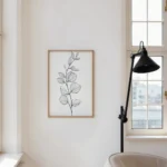 Framed black and white botanical sprig line art print, displayed on a light wall next to a large window, a black floor lamp, and an armchair in a cozy reading area.