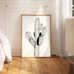 Large framed black and white line art print of a multi-stemmed cactus with black shaded areas, leaning on a wooden floor against a white wall in a bright, minimalist bedroom.
