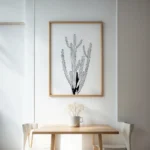 Minimalist black and white cactus art print with a light wooden frame, hanging above a simple wooden table and chairs in a bright breakfast nook.