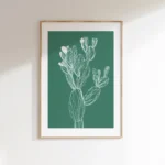 Vertical framed white line art print of a cactus on a solid green background, hung on a plain, light-colored wall with soft shadows.