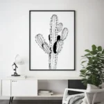 Sleek black framed black and white cactus line art print, displayed above a white console table with a desk lamp and a large green plant in a modern living room.