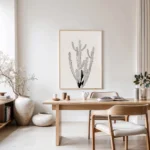 Minimalist black and white line drawing of a cactus, hanging on a light wall in a neutral home office or dining area with wooden furniture.
