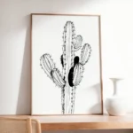 Framed black and white minimalist line art of a cactus, leaning on a light wooden dresser or shelf next to a white ceramic vase, adding a modern touch to the decor.