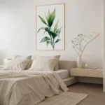 Framed art print featuring a green leafy plant branch, hung on a light wall above a bed with neutral bedding and a wooden bedside table in a serene bedroom.