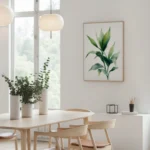 Framed art print of a green leafy plant branch, displayed on a light wall above a wooden dining table with chairs and white vases in a bright, minimalist dining space.