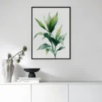 Black framed art print of a green leafy plant branch, displayed above a sleek white console table with minimalist decor and a vase in a modern living room.