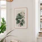 Framed art print of a Monstera plant, displayed on a wall above a wooden counter or shelf in a light, minimalist room with a large window and subtle green accents.