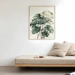 Framed art print of a Monstera plant with muted green leaves, hung above a light beige daybed or sofa in a minimalist and serene living room setting.
